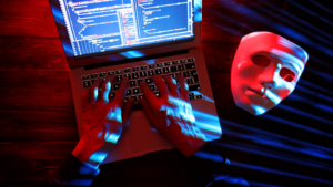 A hacker typing on a laptop with phishing codes displayed on the screen and a mask symbolizing deception, highlighting the dangers of phishing scams on social media targeting businesses.