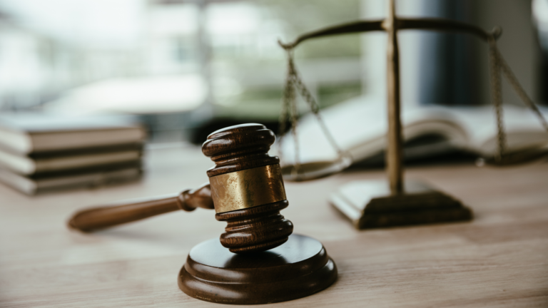A wooden judge's gavel resting on a sound block is placed on a desk, symbolizing legal authority. In the blurred background, a set of law books and a balance scale are visible, representing justice and legal certainty for investors.