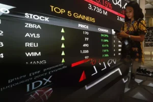 A woman checks her phone near an electronic board showing top stock gainers at the Indonesia Stock Exchange, reflecting the rising participation of Indonesian stock investors.
