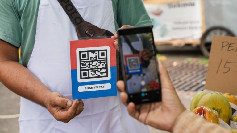 Customer using QRIS cross-border payment to scan a vendor's QR code at a local fruit market.