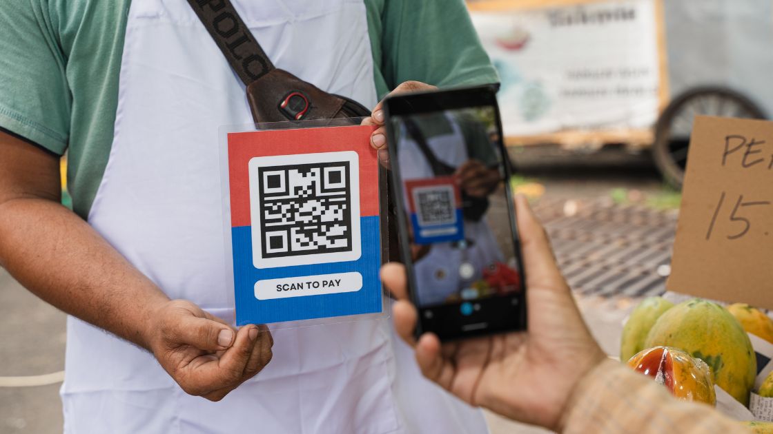 Customer using QRIS cross-border payment to scan a vendor's QR code at a local fruit market.