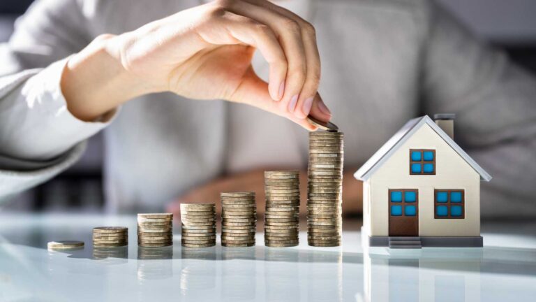 A person's hand places a coin on a growing stack of coins next to a miniature house, symbolizing investment and growth in the Indonesia housing market for foreign investors.