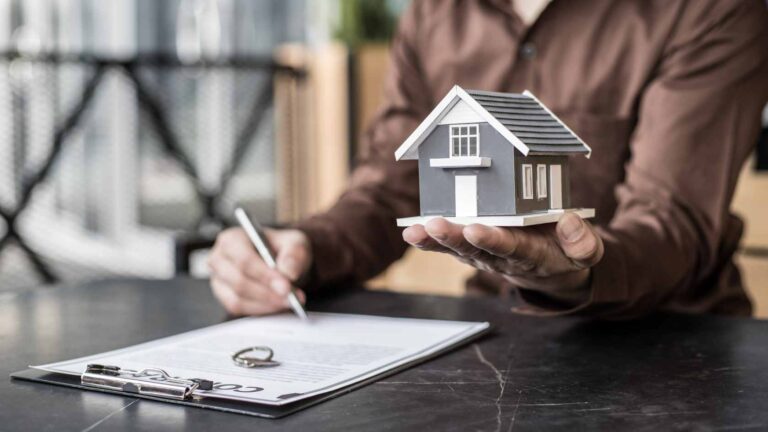Person signing a property contract while holding a miniature house, symbolizing compliance with property broker regulations.